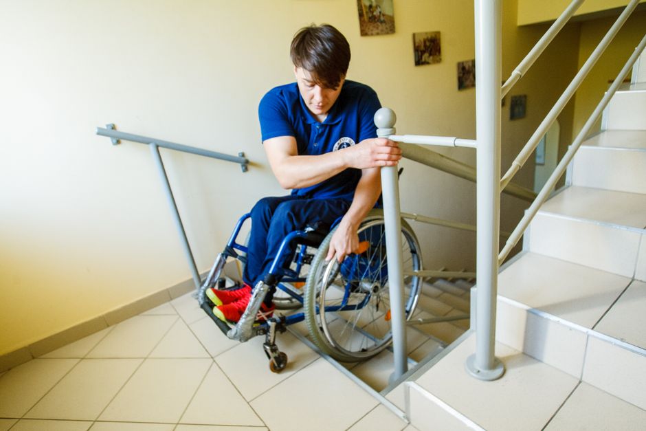 A person in a wheelchair is seen navigating down a tiled staircase with an indoor metal railing on one side. The individual is holding onto the railing with one hand while carefully managing the wheel