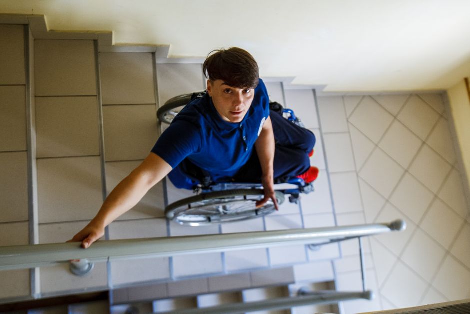 A person in a wheelchair is seen navigating down a tiled staircase with an indoor metal railing on one side. The individual is holding onto the railing with one hand while carefully managing the wheel