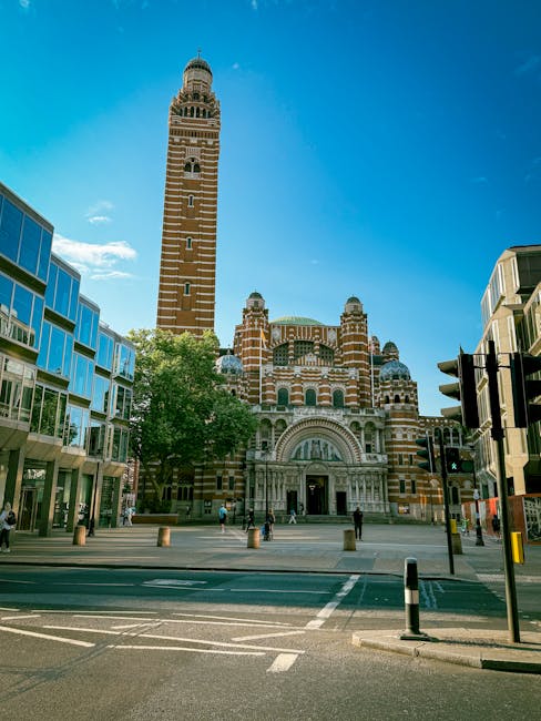 Photograph of the iconic St Mark's Campanile bell tower and the Basilica di San Marco in Venice, Italy, with clear blue sky and a few scattered clouds. The tall brick tower features a square base with arched openings near the top and a pointed spire crowned with a golden statue. In front, the basilica's ornate façade displays intricate marble detailing, arches, and domed roofs, with visitors walking along the paved square. Modern buildings with large glass windows flank the scene on either side, providing a contrast to the historic architecture. The area is bustling with pedestrians, and the street in the foreground includes a crosswalk and bollards, indicating an urban setting. The lighting is bright and natural, highlighting the vibrant colors of the brickwork and surrounding structures, capturing the essence of a typical cityscape near a major landmark, relevant to house removals or moving services in urban environments.