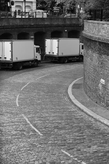 A black-and-white photograph showing two large white moving vans with closed and unmarked cargo doors parked on a curved cobblestone driveway beneath a brick archway, part of a multi-level structure with a stone wall on the right side. The vans are positioned side by side, with the van on the left partially obscured by the other, and the area around them is clear. In the background, there is a brick wall with metal railings on top, supporting trees and a building with signs on the upper level. The scene appears to be part of a home relocation operation, with the vans ready for furniture transport and packing and moving activities conducted by professionals such as Man and Van Haringey, which is relevant to the Alexandra Palace removals specialist services in Haringey.