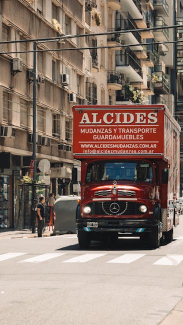 A red moving truck with a prominent advertisement for Alcides and transport services parked on a city street in Haringey. The truck is positioned in front of multiple multi-storey residential building