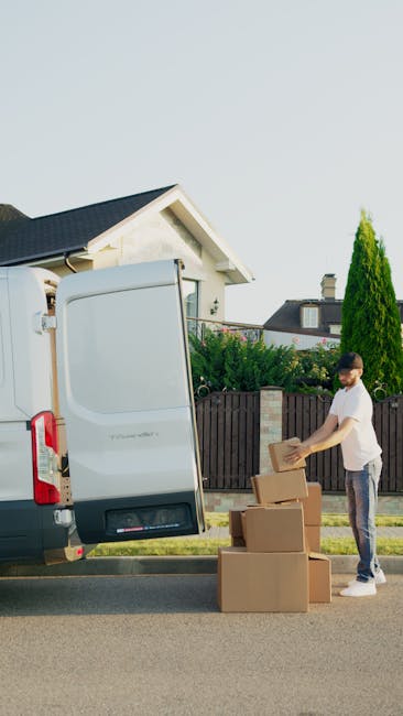 A man wearing a white t-shirt and jeans is standing next to a light grey moving van with its rear doors open, outside a residential property on a paved street. He is holding a small cardboard box, pre