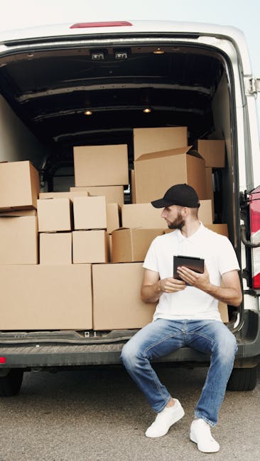 A man wearing a white t-shirt and jeans is standing next to a light grey moving van with its rear doors open, outside a residential property on a paved street. He is holding a small cardboard box, pre