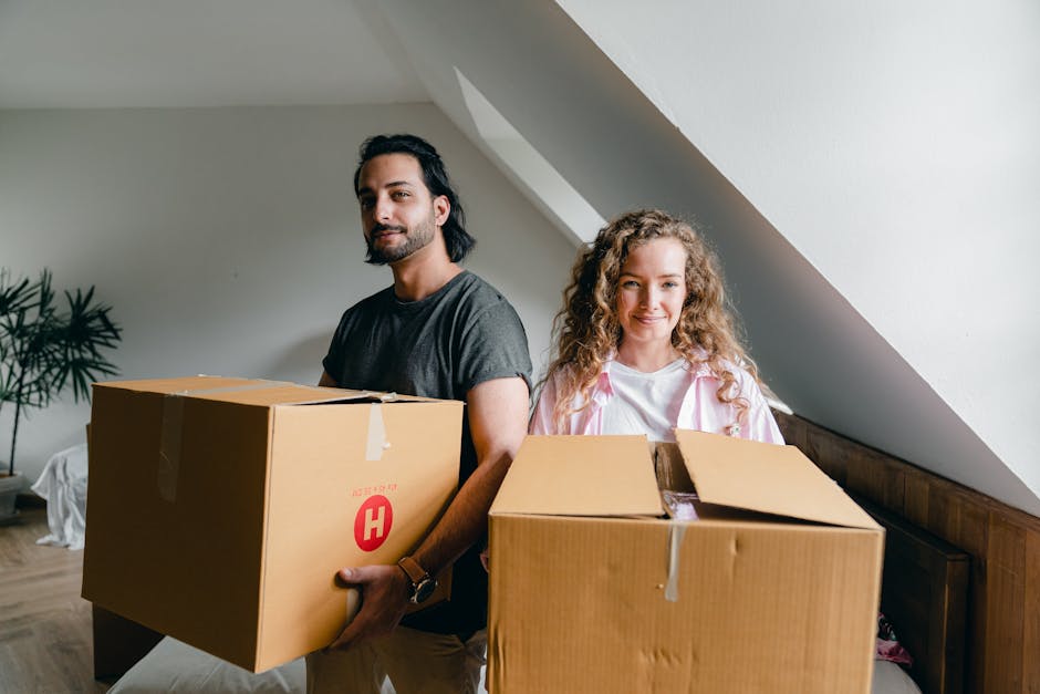 A man and a woman stand indoors in a room with a sloped ceiling and neutral walls, each holding a large cardboard moving box filled with household items. The man, with dark hair and a beard, wears a d