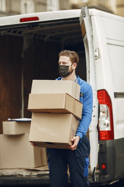 A young man wearing a black face mask, blue jacket, and dark trousers is seen standing outside at the rear of a white van during a home relocation. He is holding three cardboard boxes of varying sizes