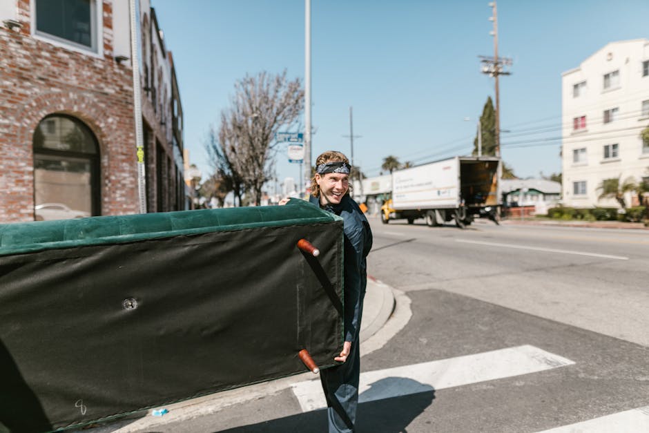 A male mover from Man and Van Haringey is standing outdoors at a street corner in Wood Green N22, during a home relocation process. He is smiling and wearing a dark jacket and a patterned headband, carrying a large, rectangular piece of furniture covered with a protective green and black moving blanket, secured with straps. The furniture is being transported on a trolley with red handles, which is positioned on the pavement. In the background, a white delivery truck is parked on the road with its back doors open, indicating loading or unloading activity. The scene includes nearby brick and white apartment buildings, leafless trees, and utility poles with wires, with bright daylight and clear skies overhead. The surroundings suggest an urban setting conducive to furniture transport and packing arrangements typical of narrow street moves in the area, and the image demonstrates careful handling of belongings during the loading process for a house removal, as managed by Man and Van Haringey.